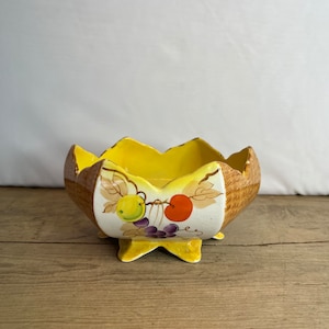 May include: A yellow ceramic bowl with a fruit design. The bowl has a scalloped edge and a woven pattern on the side. The bowl is on a wooden surface.