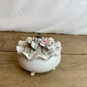 Puede incluir: Una caja de baratijas de cerámica blanca con una tapa con volantes, adornada con delicadas flores pintadas a mano en tonos rosa, azul y blanco. La caja tiene un ribete dorado y se apoya en tres pequeños pies. La tapa presenta un pequeño detalle en forma de hongo marrón.