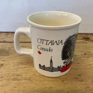 May include: White ceramic mug featuring the text "OTTAWA Canada" with a red maple leaf and a black and white illustration of a building and a person in a red coat and hat. The mug has a handle.