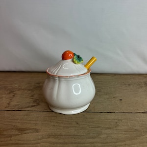 May include: White ceramic honey pot with an orange and green lid and a small yellow spoon. The pot has a decorative orange rim.