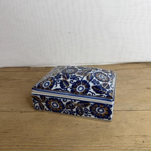 May include: A square ceramic trinket box with a lid. The box is white with a blue floral pattern and gold accents. The box is on a wooden surface.