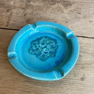 Könnte beinhalten: Ein runder, türkisfarbener Aschenbecher mit strukturierter Oberfläche. Die Mitte zeigt ein dunkleres blaues, abstraktes Design. Der Aschenbecher hat vier Kerben für Zigaretten. Der Keramik-Aschenbecher ist ein Vintage-Artikel.