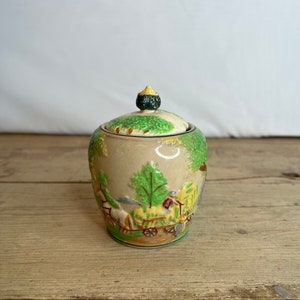 Puede incluir: Un azucarero de cerámica vintage con tapa. El cuenco es de color beige con una tapa verde y amarilla y un pequeño pomo verde. Los lados del cuenco presentan una escena pastoral con árboles, un carro tirado por caballos y figuras. El cuenco está sobre una superficie de madera.