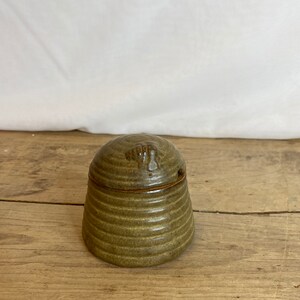 Op de afbeelding: Een kleine, handgemaakte keramische pot met een deksel. De pot heeft een gedempte olijfgroene kleur met horizontale ribbels. Het deksel heeft een klein handvat en een gat voor een lepel. De pot staat op een houten oppervlak.