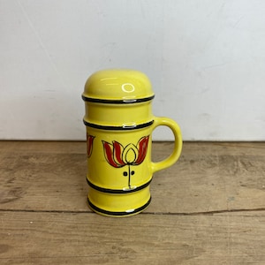 May include: A yellow ceramic beer stein with a handle and a lid. The stein is decorated with red and black floral designs. Black bands encircle the top, middle, and bottom. The background is a light-colored wall and a wooden surface.