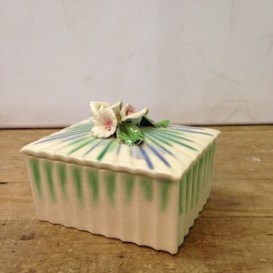 Puede incluir: Una caja de cerámica blanca con un diseño a rayas verde y azul y un acento floral. Las flores son blancas con centros rosados.