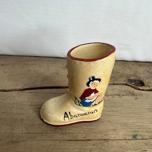 May include: A beige ceramic boot-shaped vase with a red rim and the word "Aberaeron" written on the front. The boot features a painted image of a person in traditional clothing. The vase is on a wooden surface.