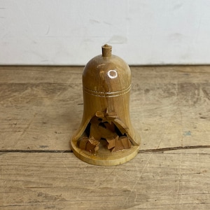 May include: A wooden bell-shaped ornament. The light brown bell features a carved design of leaves and stars at the base. It has a small, rounded knob at the top. The item is made of wood.