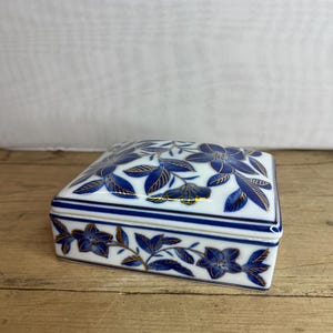 May include: A square, white ceramic trinket box with a lid, decorated with a blue floral pattern and gold accents. The box has a blue and gold floral design on the lid and sides, with a gold trim.