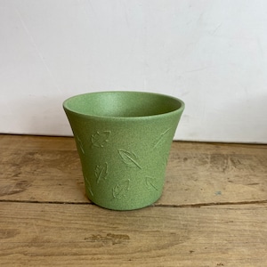 May include: A small, light green ceramic pot with a textured surface and leaf imprints. The pot has a slightly flared shape and sits on a wooden surface. Ideal for small plants or succulents.