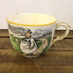 May include: A vintage ceramic teacup with a whimsical illustration. The cup features a cartoon figure in a long coat and hat, holding a mug and a walking stick. The cup has a yellow rim and a branch-like handle.