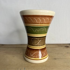 Op de afbeelding: Een decoratieve keramische vaas met een crèmekleurige boven- en onderkant. De vaas heeft horizontale banden in bruin, groen en geel met witte krul- en bladmotieven. De vaas heeft een zandloper vorm en staat op een houten oppervlak.