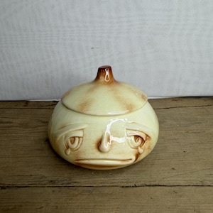 May include: A ceramic onion-shaped container with a lid. The container is light yellow with brown accents and a face design with tear drops. The lid has a small brown knob.