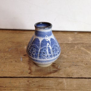 Op de afbeelding: Een kleine, handgemaakte keramische vaas met een afgeronde body en een smalle hals. De vaas heeft een blauw en wit bloemmotief rond de body en een donkerbruine rand. De vaas staat op een houten oppervlak.