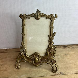 Puede incluir: Marco de fotos de latón dorado con una abertura rectangular. El marco presenta intrincados diseños florales y de volutas, con una parte superior e inferior decorativas. El marco se apoya en dos pequeños pies y tiene un respaldo de color claro. Mide unos 15 cm de alto.