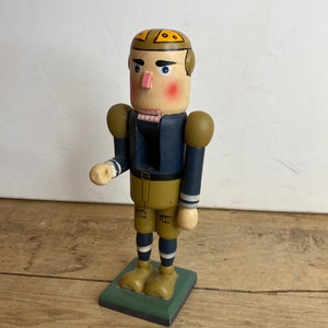 May include: Wooden nutcracker figurine depicting a football player. The figure has a painted face with rosy cheeks and blue eyes. It wears a navy blue uniform with gold details and a matching helmet. The base is green. The figurine stands on a green base.