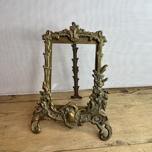Puede incluir: Marco de fotos de latón ornamentado con un diseño floral y de volutas detallado. El marco tiene una abertura rectangular y una base decorativa con patas curvas. El marco es de latón y tiene un aspecto vintage.