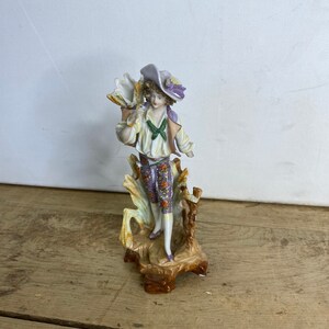 Puede incluir: Figura de porcelana de un hombre con vestimenta del siglo XVIII. La figura lleva sombrero, camisa blanca con lazo verde, abrigo morado y pantalón estampado. Montada sobre una base decorativa con diseño de árbol.