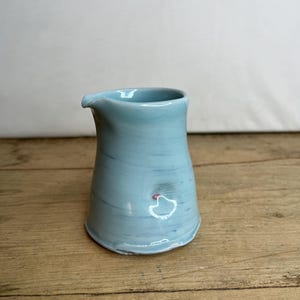 May include: A small, light blue ceramic creamer with a slightly flared spout. The creamer has a smooth, glazed finish and a subtle swirl pattern.