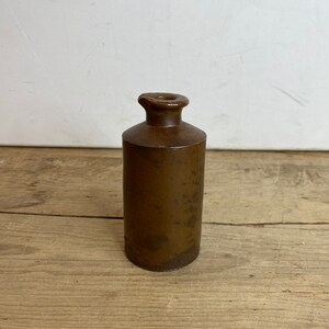 May include: A brown, cylindrical stoneware bottle with a narrow neck and a small, rounded opening. The bottle has a dark, matte finish and appears to be antique. The bottle is approximately 6 inches tall.
