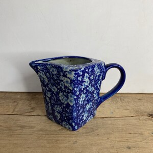 May include: A blue and white floral patterned jug with a handle. The jug has a unique angular shape and a spout for pouring. The floral pattern is a delicate design of white flowers on a blue background. The jug is likely made of ceramic.