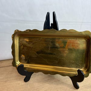 May include: A gold-coloured metal tray with a scalloped edge. The tray has an engraved map of The Garden Isle, which is another name for Kauai, Hawaii. The tray is on a black stand.