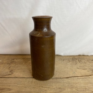 May include: A brown stoneware vase with a cylindrical body and a slightly flared neck. The vase has a rustic, aged appearance, with variations in the brown color. The vase is displayed on a wooden surface.