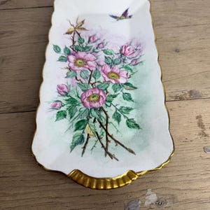 May include: A rectangular, off-white decorative tray with a gold scalloped edge. The tray features a floral design with pink roses, green leaves, and a small blue bird. The tray is likely made of porcelain or ceramic.