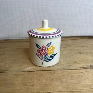 Puede incluir: Un pequeño tarro de cerámica blanquecina con tapa. El tarro está decorado con un diseño floral en rojo, amarillo y azul. La tapa tiene un patrón de rayas amarillas y moradas. El tarro está sobre una superficie de madera.