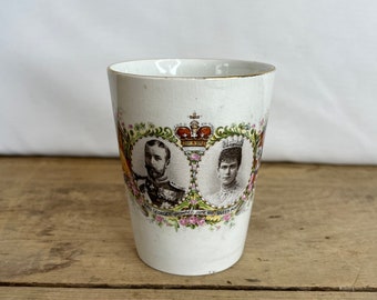 Puede incluir: Taza conmemorativa de cerámica blanca con borde dorado. La taza presenta retratos del rey Jorge V y la reina María, rodeados de diseños florales y coronas. El texto en la taza dice "King George V Crowned June 1911 Queen Mary".