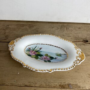 Peut inclure: Plat décoratif ovale blanc avec une bordure dorée et une scène peinte de nénuphars. Le plat a un bord festonné et des accents dorés sur les coins. La peinture de nénuphars présente des fleurs roses et des nénuphars verts.