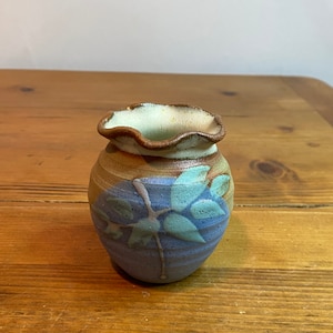Puede incluir: Un pequeño jarrón de cerámica hecho a mano con un borde ondulado. El jarrón presenta un esquema de color azul y marrón con un diseño floral. El jarrón mide aproximadamente 10 cm de alto y 8 cm de ancho.