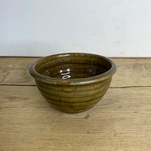 May include: An olive-green ceramic bowl with a rounded shape and a slightly flared rim. The bowl has visible horizontal ridges and a glossy finish, suggesting a rustic, artisanal aesthetic. The bowl is sitting on a wooden surface.