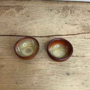 Pode incluir: Duas pequenas tigelas redondas de cerâmica com borda marrom e interior amarelo claro. As tigelas são colocadas lado a lado sobre uma superfície de madeira. As tigelas parecem ser feitas à mão e têm uma estética rústica.