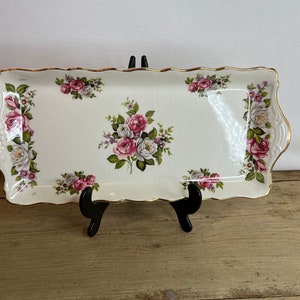 May include: A rectangular, off-white ceramic serving tray with a scalloped edge and gold trim. The tray is decorated with clusters of pink, white, and purple roses. The tray is supported by a black stand.