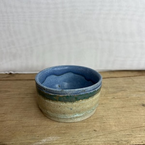 May include: A small, handmade ceramic bowl with a rounded shape. The interior is a light blue, with a darker blue rim. The exterior features bands of green, tan, and cream colours. The bowl sits on a wooden surface.