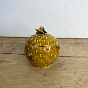 Puede incluir: Un tarro de miel de cerámica con un diseño de cesta tejida en tonos marrones y amarillos. La tapa presenta un asa con forma de abeja. El tarro tiene forma redonda y es adecuado para guardar miel u otros objetos pequeños.
