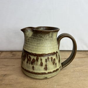 May include: A small, brown and green ceramic pitcher with a handle. The pitcher has a speckled glaze and a brown band around the middle.