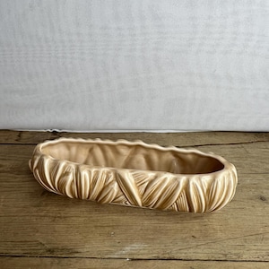 Pode incluir: Um vaso de cerâmica bege com um design texturizado e com nervuras. O vaso retangular tem uma borda superior ondulada e assenta sobre uma superfície de madeira. Adequado para uso interno ou externo.