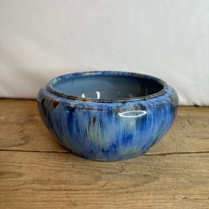 May include: A round, blue ceramic bowl with a glossy finish. The bowl has a dark blue interior and a lighter blue exterior with streaks of brown and white. The bowl sits on a wooden surface.