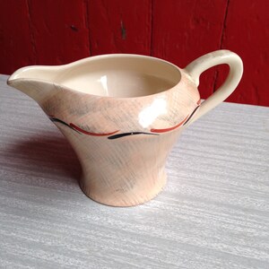 Puede incluir: Una jarra de crema de cerámica de color crema con un diseño geométrico marrón y rojo. La jarra tiene un asa y un pico.