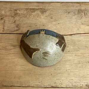 May include: A handmade ceramic bowl with a unique design. The bowl has a light beige speckled interior, a dark blue rim, and brown accents. The design includes a castle and stylized hand-like shapes. The bowl is on a wooden surface.