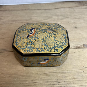 May include: An octagonal decorative box with a black rim and lid. The lid features a blue and gold floral pattern with small birds. The box is on a wooden surface.
