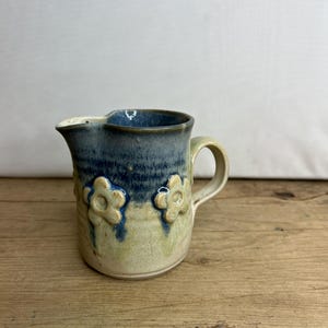 Op de afbeelding: Een kleine, blauwe en witte keramische kan met een handvat en drie reliëfbloemen. De kan staat op een houten oppervlak.