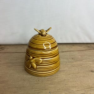 Könnte beinhalten: Keramik-Honigtopf in Bienenwabenform mit Deckel. Der Topf hat eine warme braune Farbe mit horizontalen Rillen und einer Bienenfigur oben. Eine weitere Biene befindet sich an der Seite. Der Topf steht auf einer Holzoberfläche.