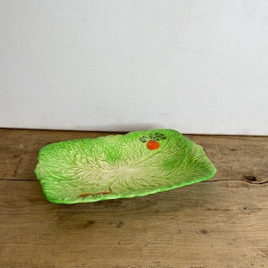 May include: A rectangular, green ceramic serving dish shaped like a head of lettuce. The dish has a textured surface and a small orange fruit design. The dish is on a wooden surface.