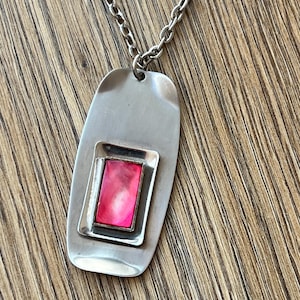 Puede incluir: Un collar con colgante plateado con una incrustación rosa rectangular dentro de un marco rectangular. El colgante cuelga de una delicada cadena. El collar se exhibe sobre una superficie de madera.