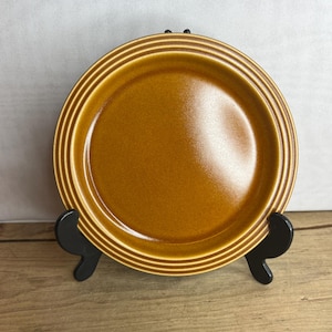 May include: A round, amber-colored ceramic plate with concentric ridged detailing around the rim. The plate is displayed on a black stand against a neutral background. The plate's diameter is approximately 9 inches.