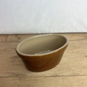 Puede incluir: Un pequeño cuenco ovalado de cerámica marrón con interior beige claro. El cuenco tiene un acabado brillante y está sobre una superficie de madera. Este cuenco es adecuado para servir salsas o pequeñas porciones de comida.