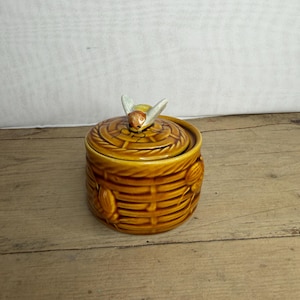 Op de afbeelding: Een keramische honingpot met een deksel, met een bijvormige handgreep. De pot heeft een warme geelbruine kleur, met een textuur, geweven mandontwerp. De bij heeft witte vleugels en een geelbruin lichaam.